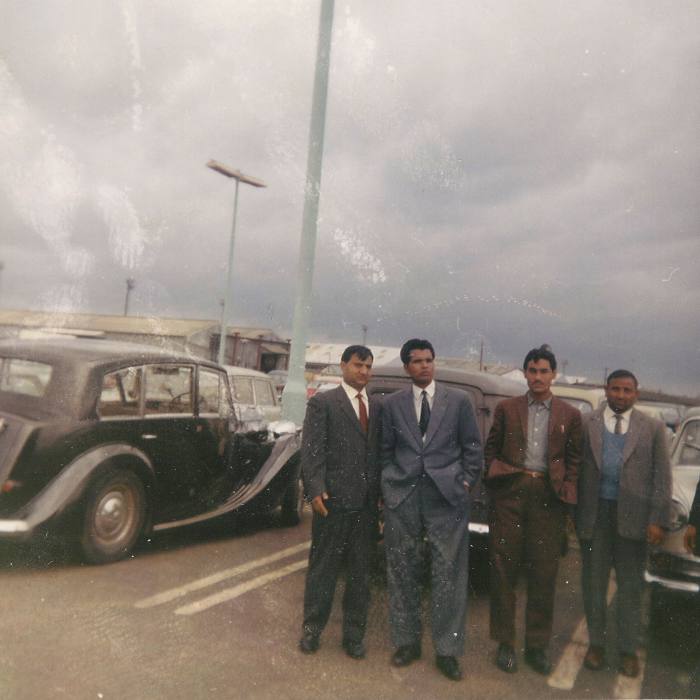 Four young men in suits pose for a photograph in 1963, standing next to a large parked car