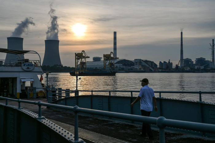 A person wearing a mask and cap walks on a pier at dusk, with an industrial landscape featuring cooling towers emitting steam, cranes, and factory buildings silhouetted against the setting sun in the background
