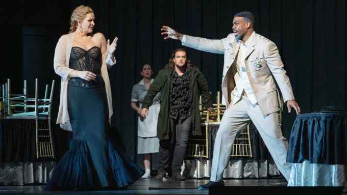 In a room with chairs upturned on tables, a man wearing a military-style jacket reaches out aggressively to a scared-looking, glamorously dressed woman; in the background another man rushes forward and a woman looks on