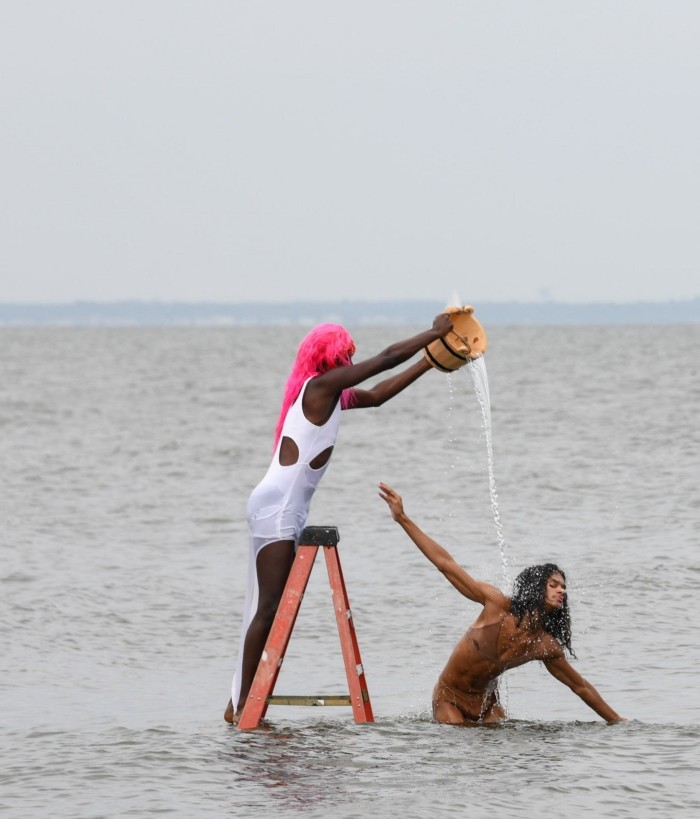 A person in a pink wig stands on a ladder in the sea pouring water over anoher person