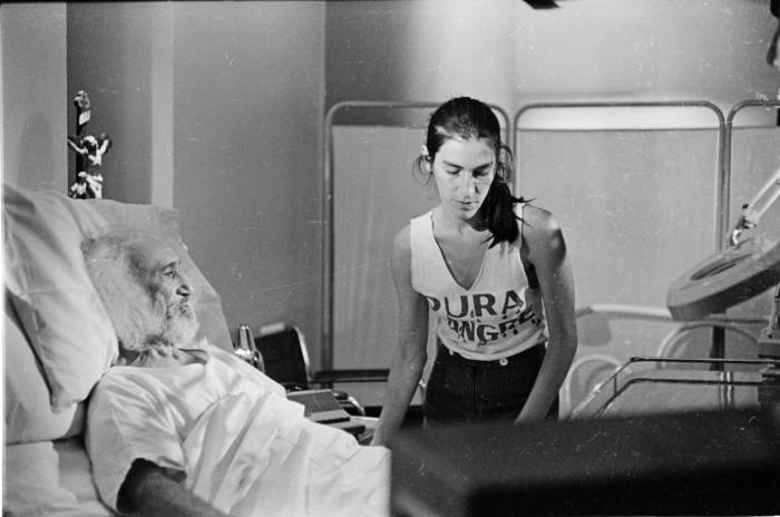 Black and white photo of Karen Lamassonne in a white sleeveless top at the hospital bedside of an old man