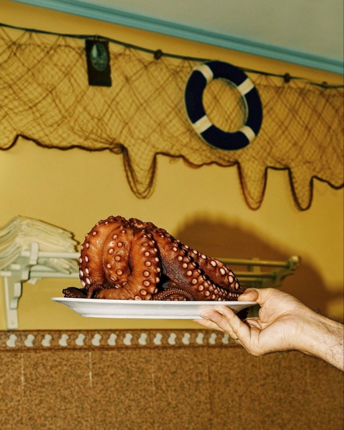 Octopus on a plate held up by a hand against a yellow wall at Ribeira do Miño