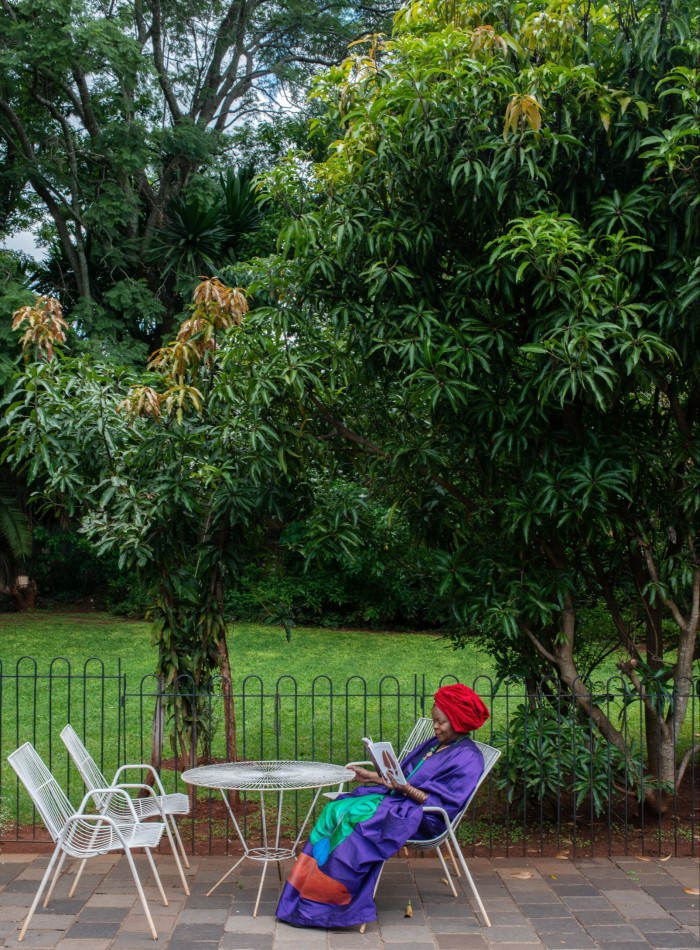 A woman in long robe and red turban sits reading at a garden table, with trees behind her