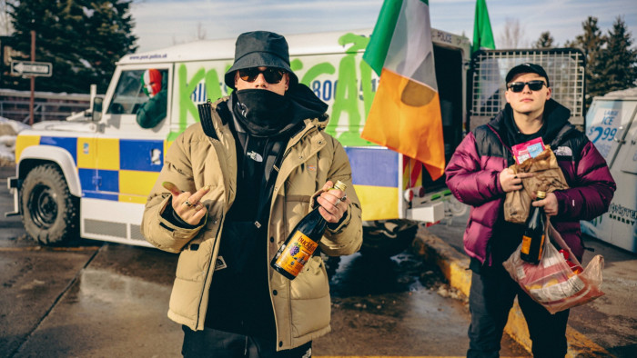 Two men stand in front of a van with the word ‘kneecap’ spray-painted along the side and decorated with Irish flags. A man wearing a mask with the colours of the Irish flag sits in the driver’s seat. Two men in sunglasses and puffer jackets stand in front holding bottles and bags of chips. One of the men gives the middle finger. 