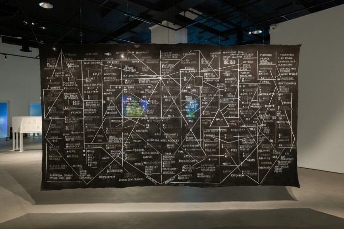 A large black canvas hanging from the ceiling of a gallery, covered in a mind map of words relating to the opium trade 