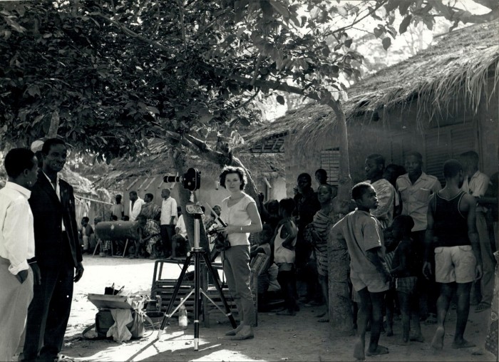 Black and white photo of a woman with a cine camera among people by a thatched hut