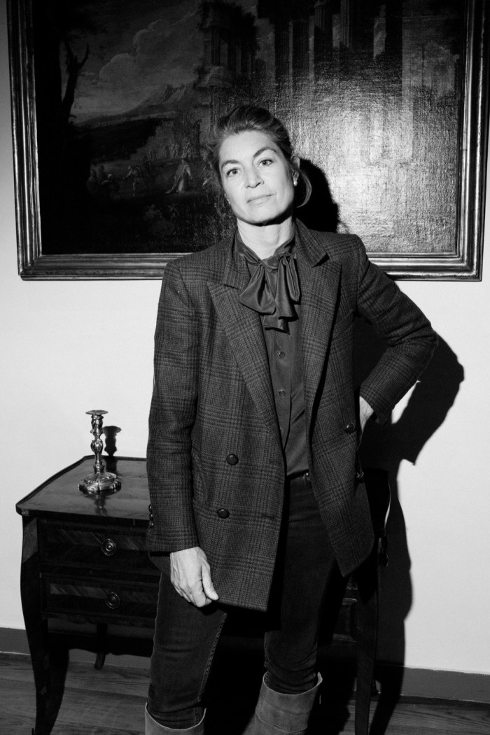Black and white photo of a woman in a heavy tweed jacket standing with one hand on her hip in front of an old painting