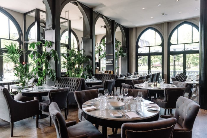 The dining space in Nimb Brasserie, with grey velvet chairs, marble tabletops, Moorish-style arches and windows and a row of tall plants