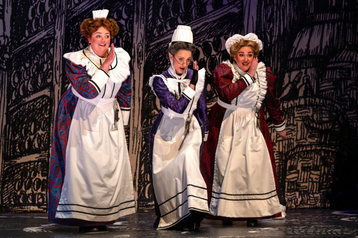 Three women clownishly made up as old-fashioned hotel maids in white aprons sing behind their hands