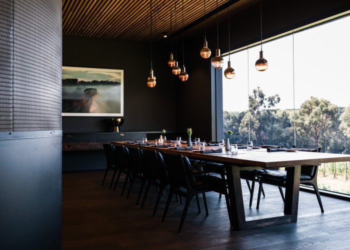 A dining room at Ten Minutes by Tractor, with a long dark-wood table, bronze global pendant lamps and floor to ceiling windows overlooking vines and gum trees