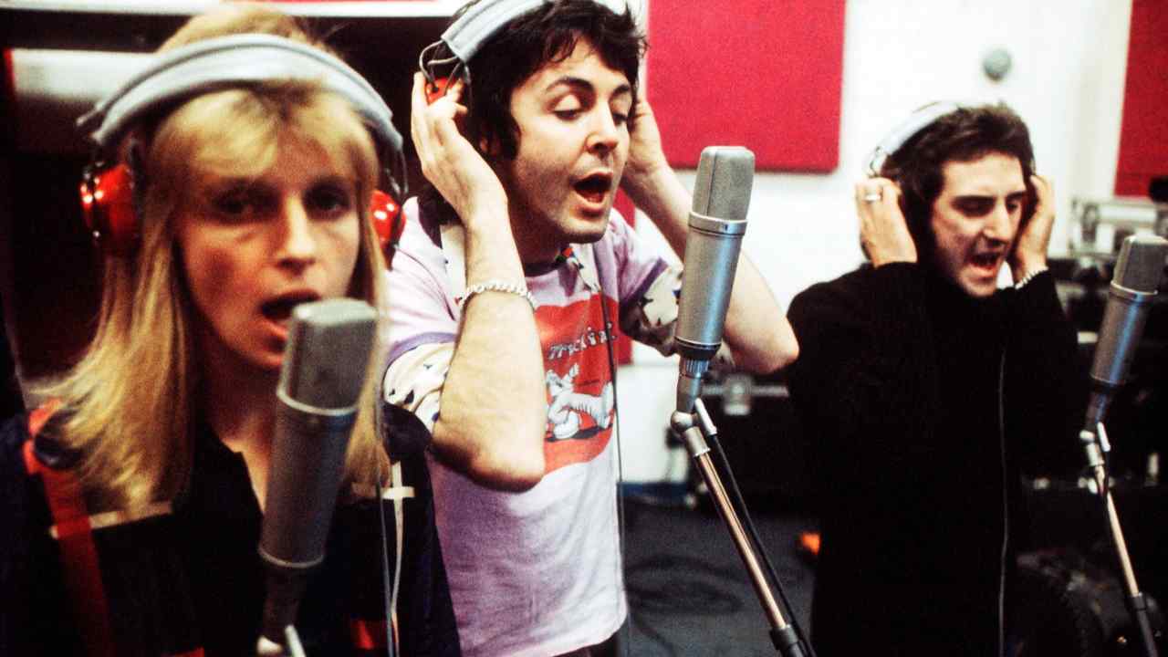 A woman and two men wearing headphones stand in a recording studio singing into microphones