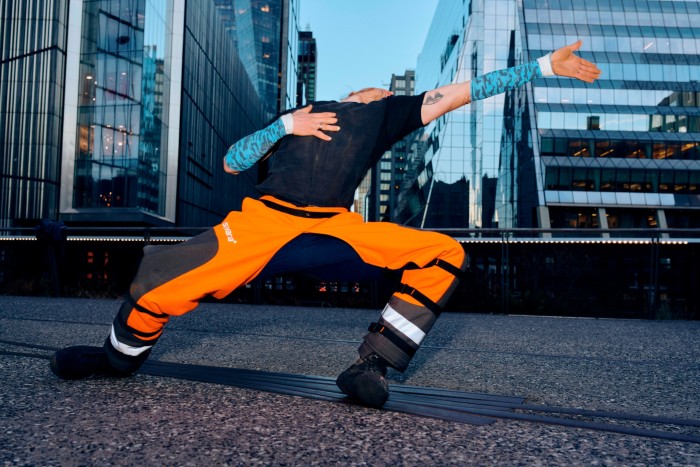 A young man wearing hi vis orange trousers and a black T-shirt is captured dancing in a city at dusk