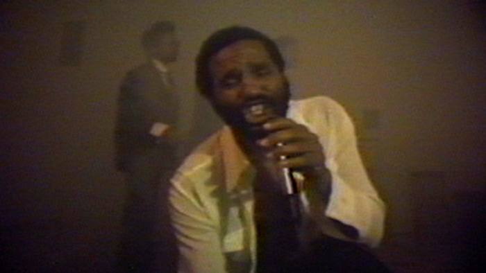 Blurry still of a man singing with a microphone to the camera