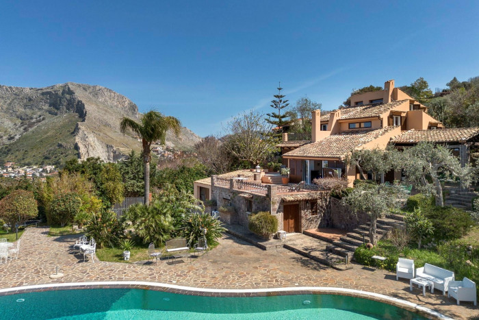 A pink-hued villa with olive trees, palm trees and a pool, surrounded by rugged countryside
