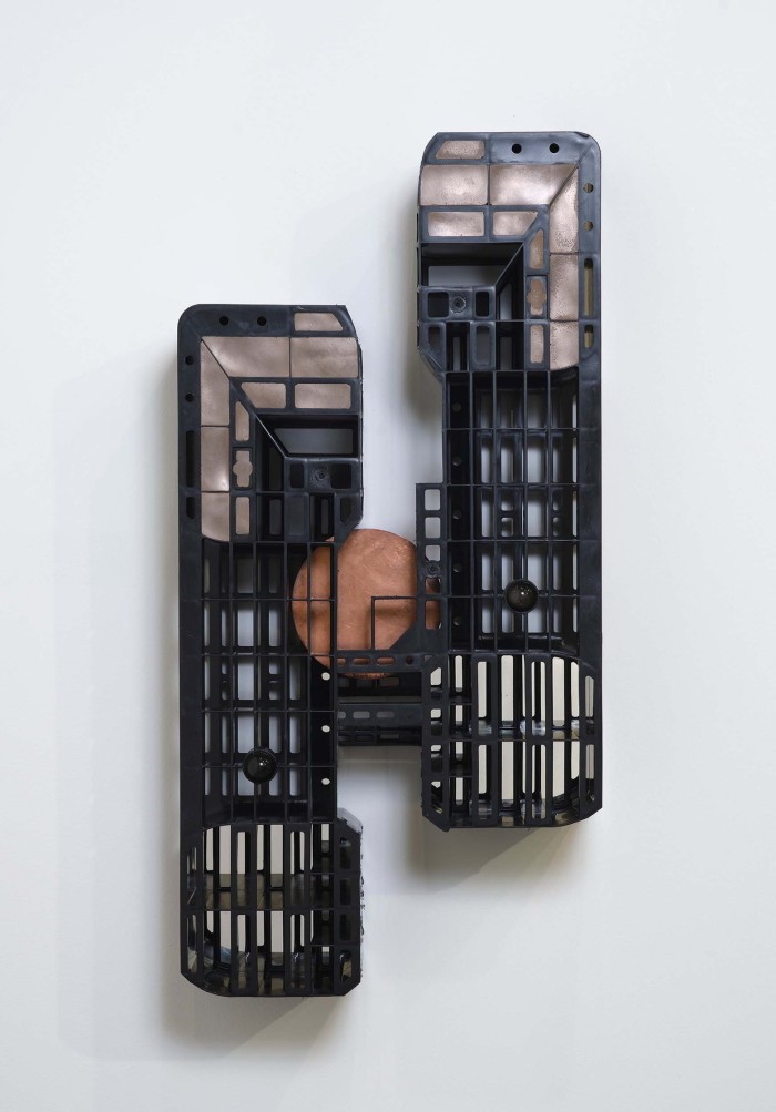 A wall-mounted sculpture featuring a bronze disk surrounded by two grid-like plastic pallet structures 