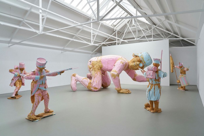 A large scultpural installation of pink-coated soldiers aiming their guns at a huge wooden figure on its hands and knees 