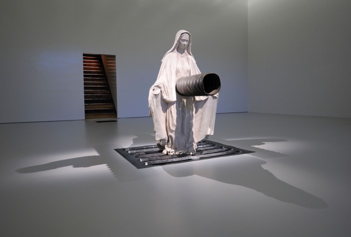A large plaster statue of the Virgin Mary, in a robe with her hands outstretched, pierced with a large pipe through the stomach