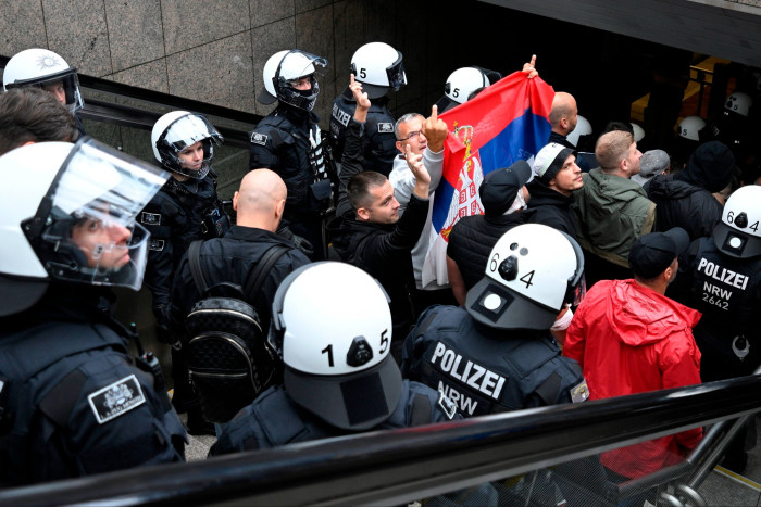 Police in white helmets walk down stairs on either side of a row of men, some of whom hold up one finger. One man holds aloft a Serbian flag 