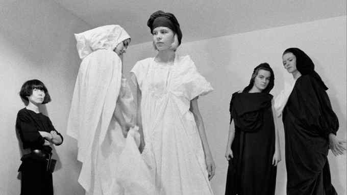 Three women wearing black outfits watch two others in all-white dresses in a studio setting