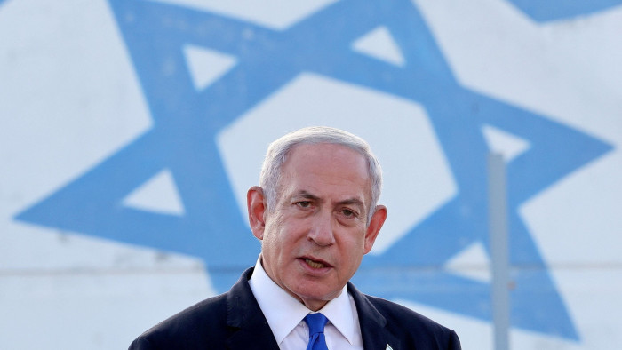 A man in a suit stands in front of a large Israeli flag