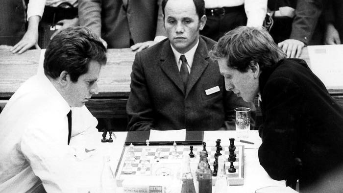 A black-and-white photograph from 1970 shows Boris Spassky on the left, in white shirt and tie, studying the chessboard and sitting opposite Bobby Fischer, wearing a dark jacket and leaning over the board