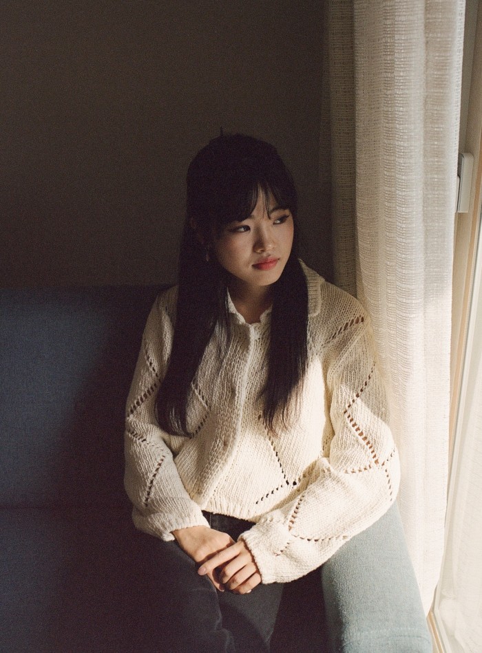 A portrait of Kim Joo Kyung sitting by a window, looking out with a wistful expression, her hands clasped on her lap. A white curtain above her left shoulder partially obscures the light  