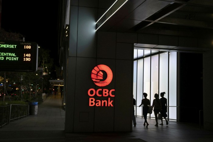 Pedestrians pass in front of Oversea-Chinese Banking Corp. (OCBC) signage illuminated at night in the shopping district of Singapore