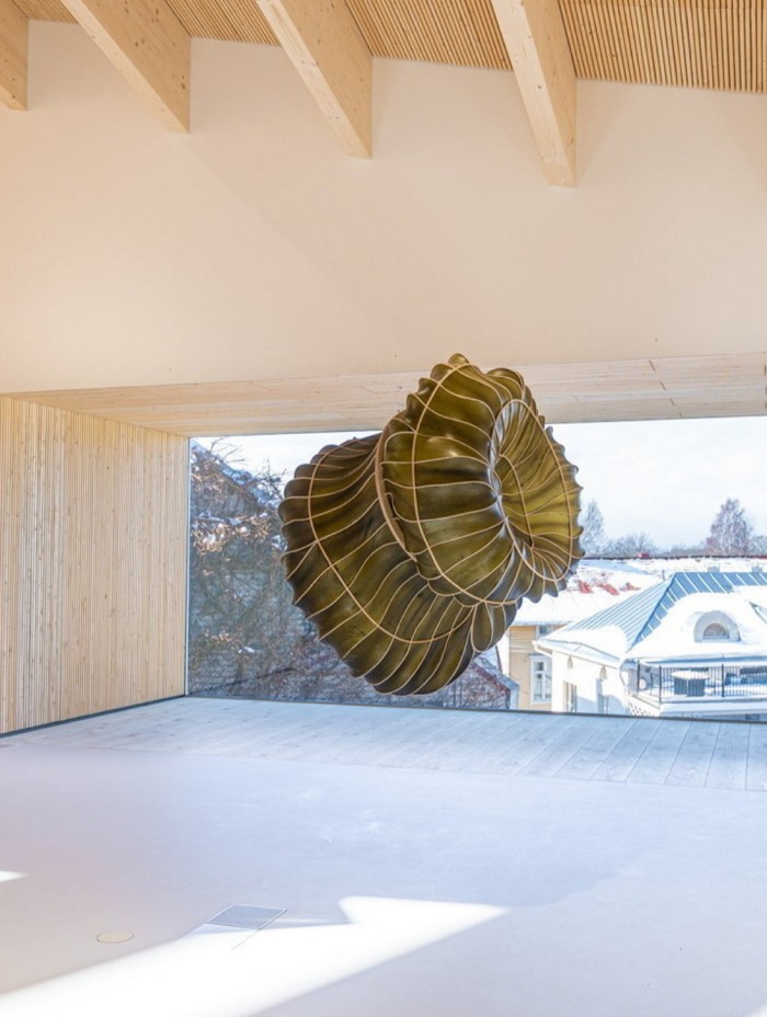 A large sculpture which looks like the neck of a bottle of wine folding in on itself and made from seaweed is suspended in a gallery 