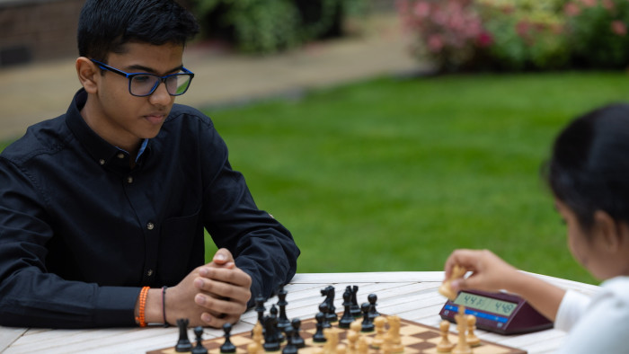 A teenage boy in dark shirt and glasses sits in front of a chessboard, his hands clasped in front of him, as his opponent, a dark-haired girl in white shirt, picks up a chess piece