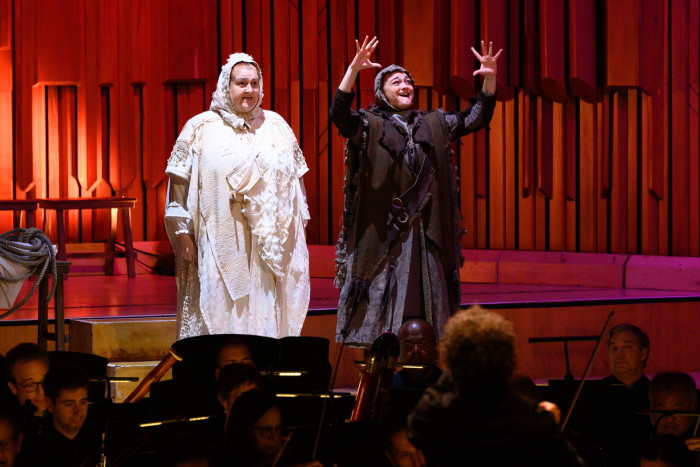 Two performers stand on stage wearing long robes; one holds her hands aloft in a gesture of wonder