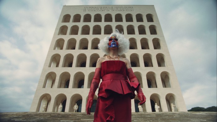 A person, seen from a low angle in front of a tall building, wearing a red ball gown