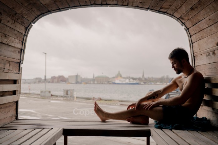 A topless man sitting beside the window at the far end of CopenHot’s Panoramic Sauna 