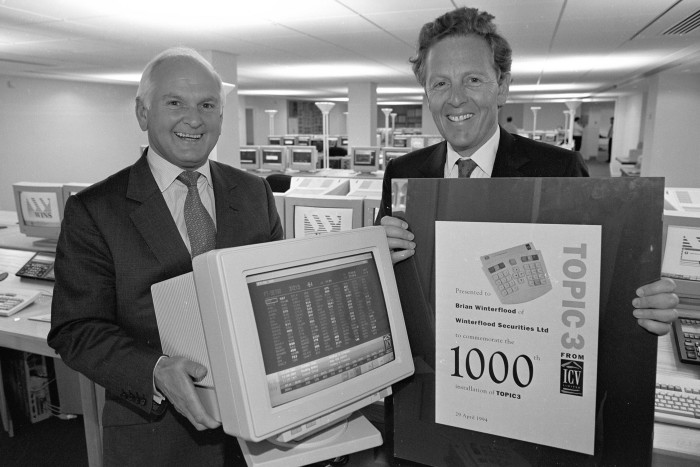 Two men in suits pose for a photo, one standing behind a 1980s computer VDU and the other holding an advertising poster