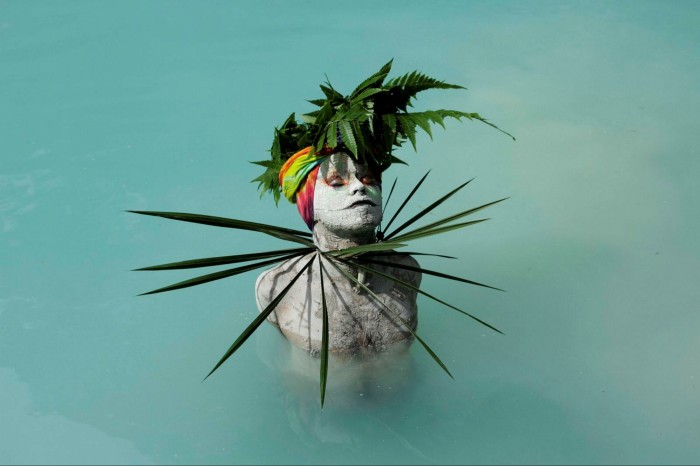 A person covered in white mud with ferns on their head walks through blue water