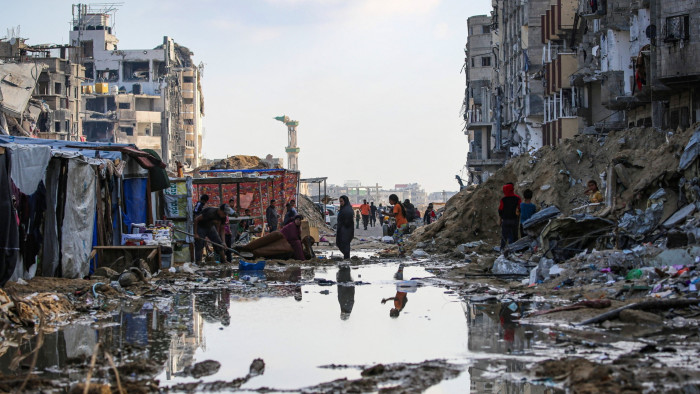 Displaced Palestinians walk around a puddle in front of destroyed buildings