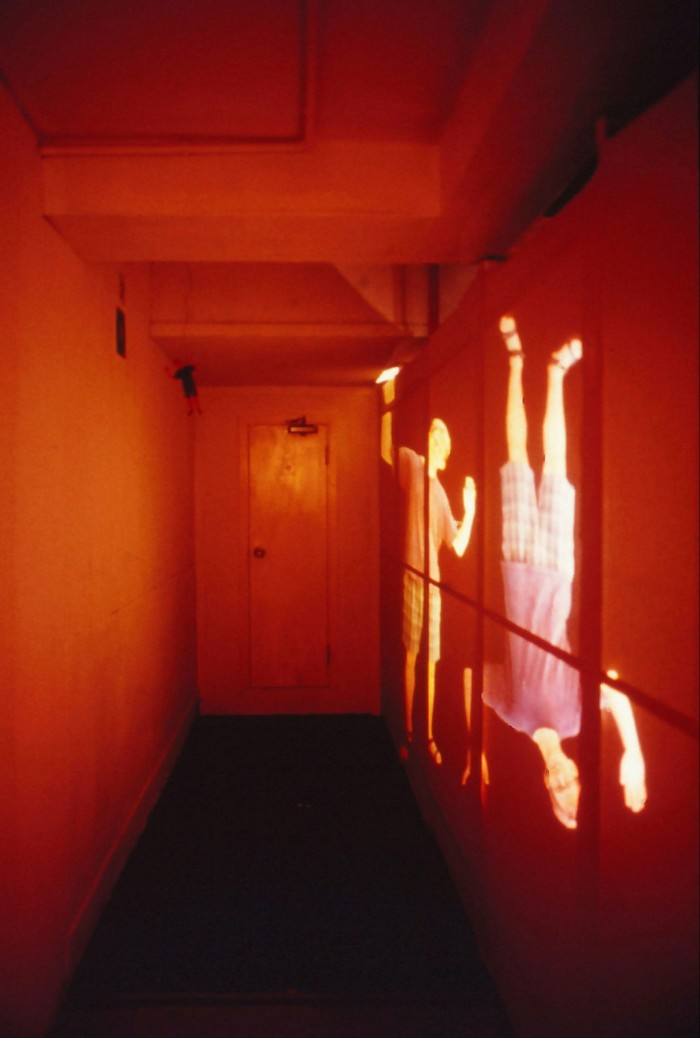 A narrow corridor is lit in a red hue with two glowing figures projected against the wall