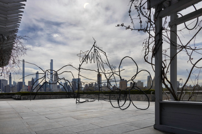 Large wiry metal sculptures, including two in the shape of a spider and a flower, are placed on the flat roof of a building with a skyline of skyscrapers in the background