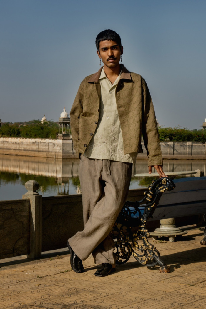 A model stands on a dock, leaning against a bench. He wears a loose jacket over an embroidered shirt and loose trousers