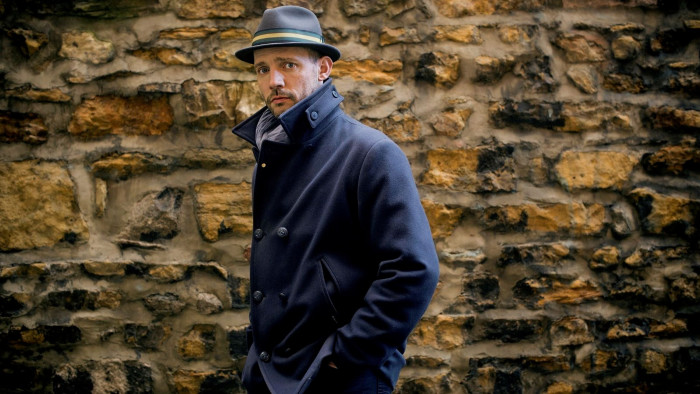 A man in a fedora and navy pea coat walking in front of a rough hewn wall