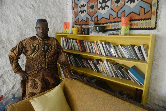 Writer Binyavanga Wainaina stands in the corner of a room in between a bookcase full of books and a sofa 