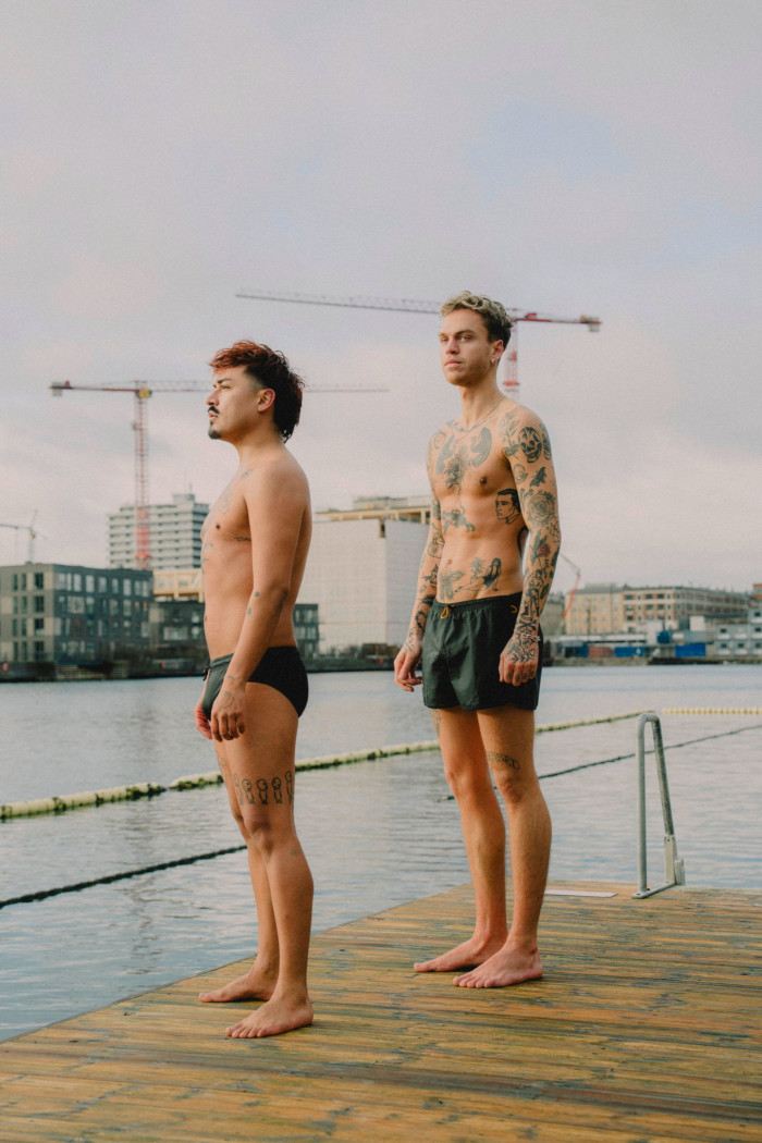 Two men, one heavily tattooed, stand on a deck near water, one is sports briefs, one in longer swim trunks