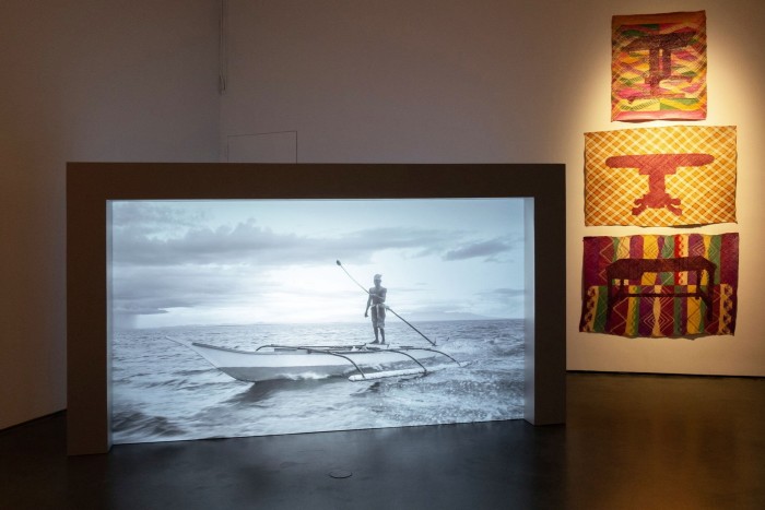 On the left, a large black and white video still of a man in a canoe. On the right, fabric pictures in red, green and yellow on the wall