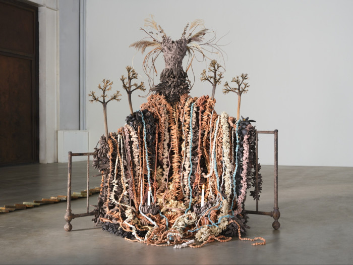 An installation incorporating textiles, natural fibres, coral beads and plants creates a humanoid silhouette in tones of brown, pink, light blue and white