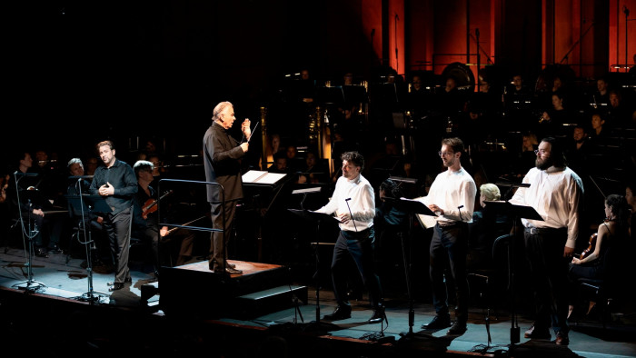 Four male singers performing on stage in front of an orchestra and conductor, wearing black