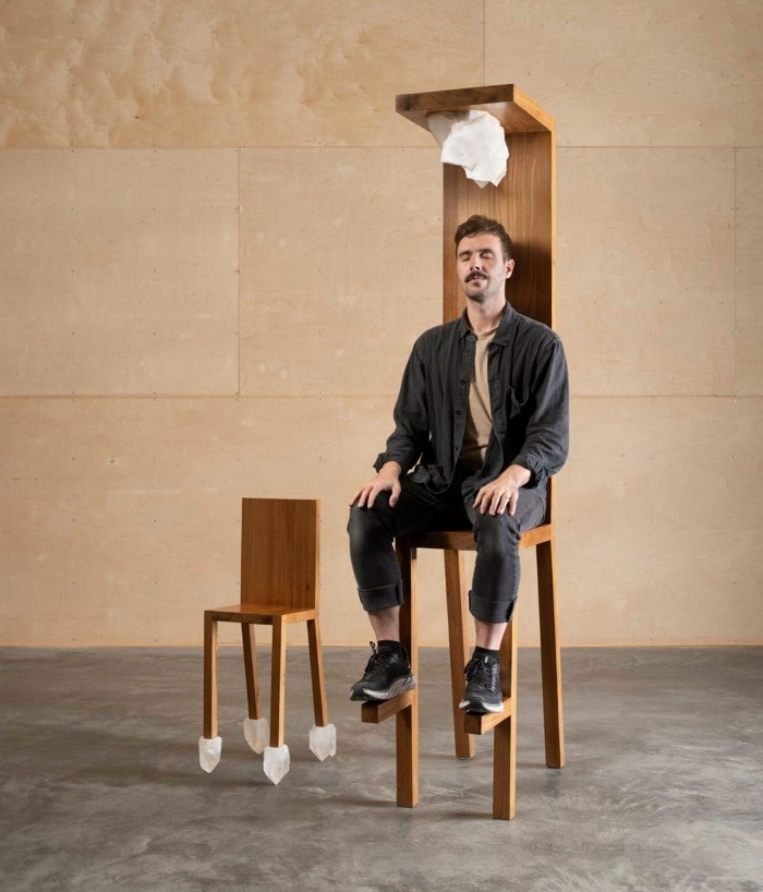A man sits in an absurdly high wooden chair with a mini-ceiling from which hangs a large white crystal over his head