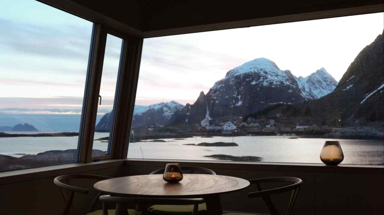 A hotel room’s wide windows look out across the sea to jagged snowy mountains