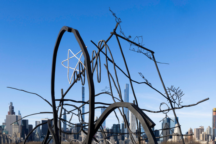 Wiry metal sculptures are seen against a blue sky and a skyline of tall buildings