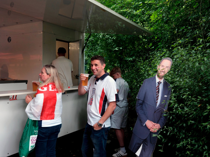 A woman and a man in England shirts stand by a food van. The man, smiling, holds up a glass of beer. A young boy is behind them, and on the right is a cardboard figure of a smiling man in suit and tie