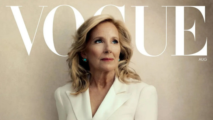 A smiling blonde woman in a white coat dress with the words ‘Vogue’ in capital letters behind her