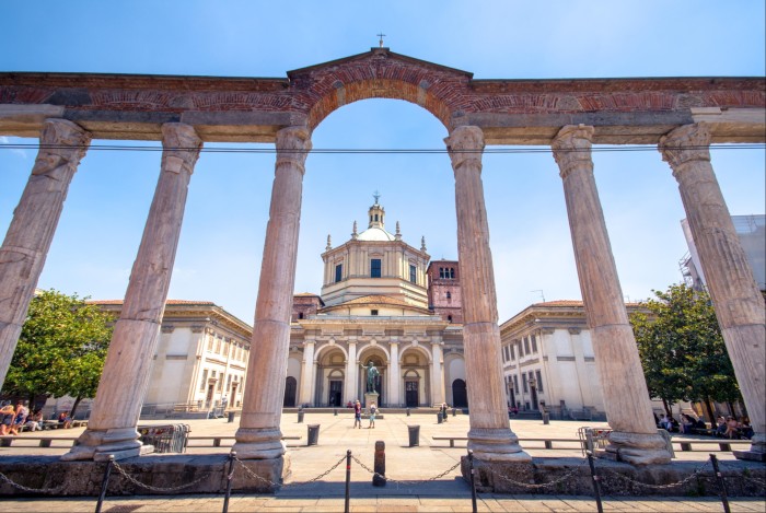 The Colonne di San Lorenzo, a Roman colonnade, beneath a blue sky, with a statue of Roman emperor Constantine and church seen through its main arch